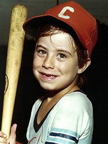 adam holds a baseball bat and smiles, top two front teeth missing. wearing red baseball cap