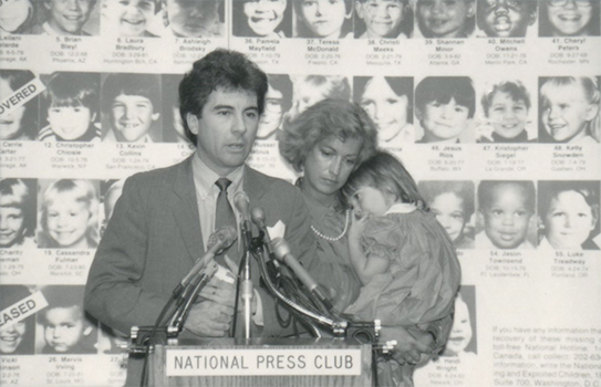 black and white: john and reve walsh speak at a podium in front of missing children posters